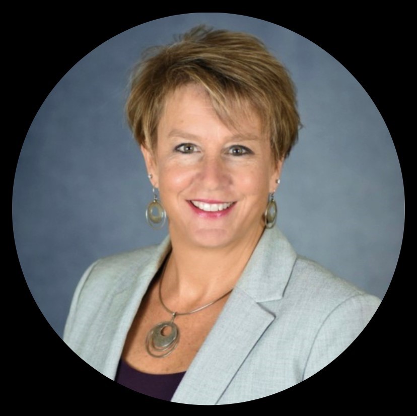 A headshot of a smiling woman with short brown hair wearing a gray blazer in a dark gray background