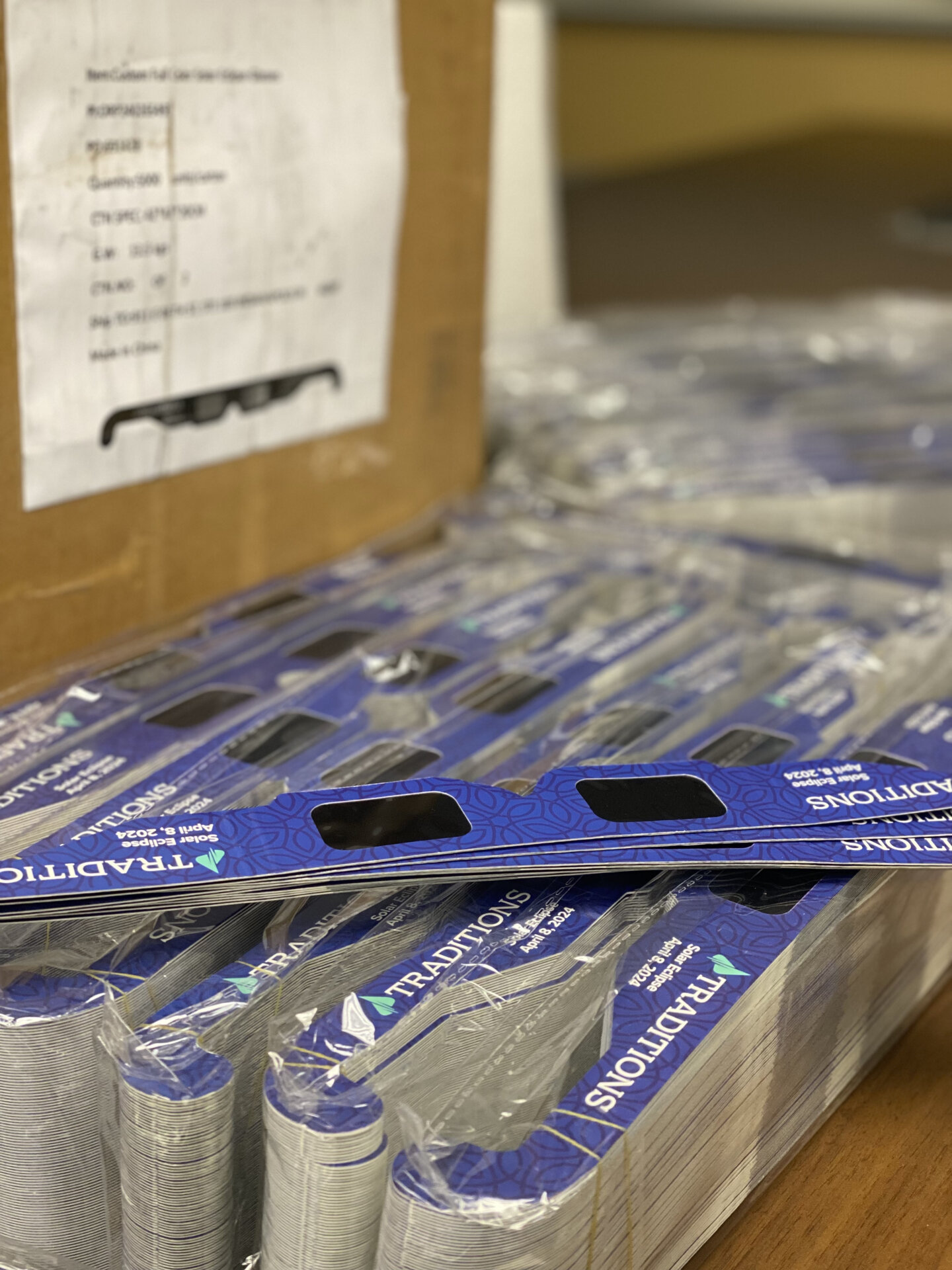 Packs of blue eclipse glasses next to a medium-sized cardboard box with white paper detailing info of glasses glued on it