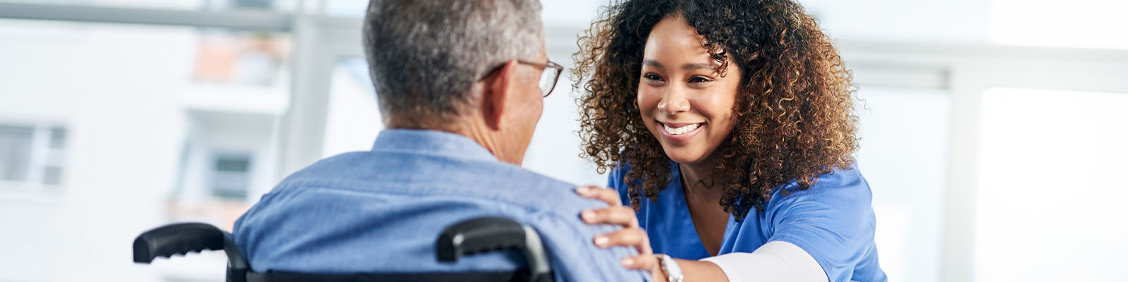 A senior man sitting in a wheelchair looking up at his nursing assistant while she smiles at him